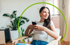 A woman sitting down using her smartphone and smiling off into the distance.\