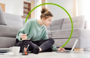 A female student sitting on the floor of her apartment with her laptop taking notes from an online college class